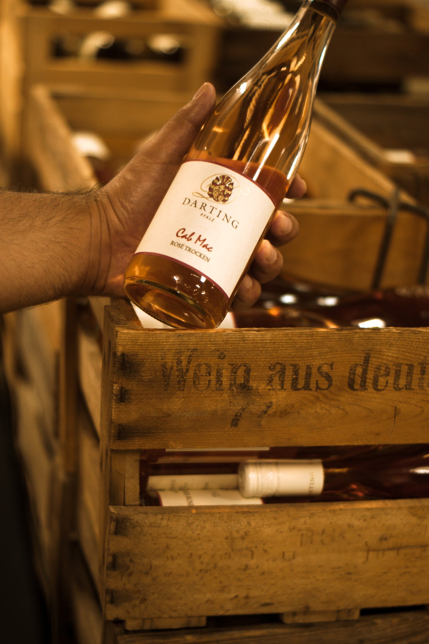 Una botella de vino rosado se sostiene con la mano izquierda y se inclina en diagonal hacia la derecha. Al fondo hay una caja de vino con muchas botellas de vino rosado.